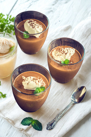 Three individual glass bowls filled with decadent chocolate mousse, garnished with whipped cream and fresh mint leaves. A spoon rests nearby.の写真素材