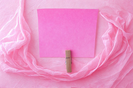 A flat lay composition featuring a square pink card held by a wooden clothespin, surrounded by flowing pink fabric.の写真素材
