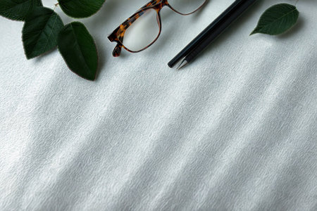 A sophisticated arrangement featuring reading glasses, a sleek pen, and lush green leaves on a textured white surface.の写真素材