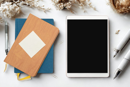 A clean and organized workspace featuring a notebook, tablet, pens, and delicate white flowers on a white surface.の写真素材