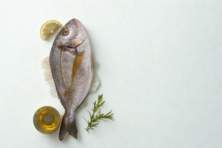 A whole, uncooked fish is presented with lemon slices and fresh herbs, ready for preparation. The composition is clean and minimalist.の写真素材