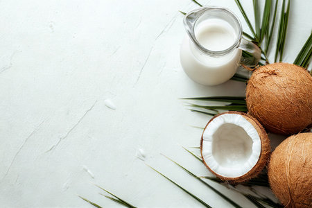 Whole and halved coconuts sit beside a pitcher of coconut milk and palm leaves on a light marble background.の写真素材