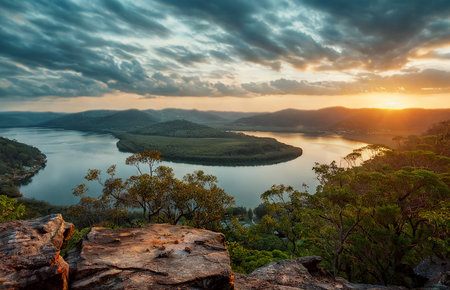 A breathtaking sunset paints the sky with vibrant colors over a serene river valley, viewed from a rocky, tree-lined overlook.の写真素材
