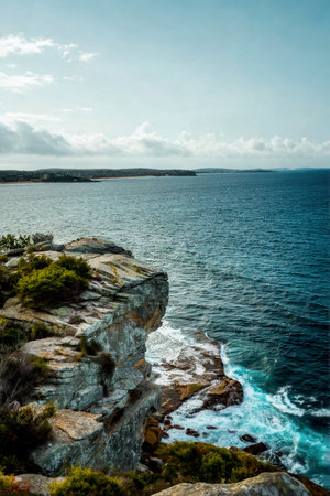 A dramatic view of weathered rock formations and green vegetation overlooking a vast, shimmering blue ocean with waves crashing against the shore.の写真素材
