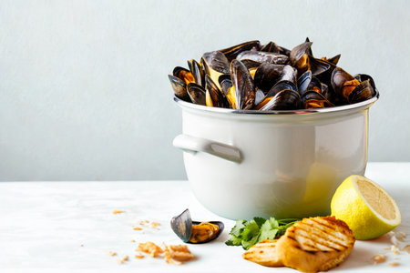 A large metal bucket overflowing with freshly steamed mussels, served with grilled bread and lemon wedges on a white surface.の写真素材