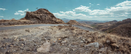 A winding road carves through a rugged, dry mountainous terrain with sparse vegetation and a dramatic rock formation under a bright blue sky.の写真素材