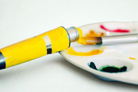 A close-up shot of a yellow paint tube next to a white art palette with various colorful paint smudges and a brush.の写真素材