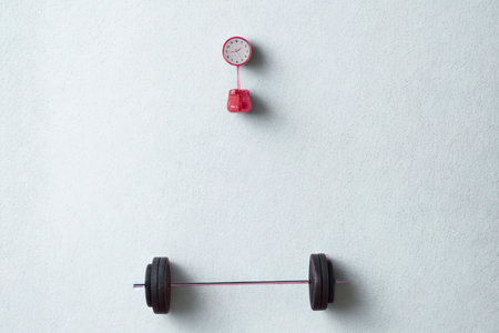 A barbell rests below a hanging scale with a red object, symbolizing strength training and progress tracking.の写真素材