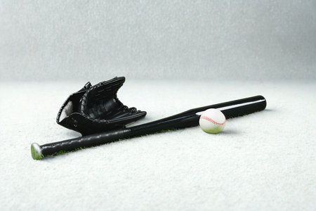 A black baseball bat, glove, and ball are artfully arranged on a plain, light-colored surface.の写真素材