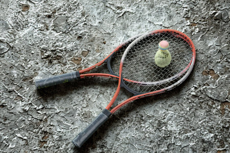 A pair of red and gray badminton rackets lie crossed on a textured, gravelly surface with a single shuttlecock resting in one of the nets.の写真素材