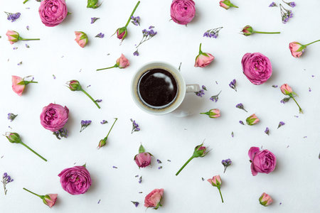 Overhead view of a white coffee cup filled with dark coffee, artfully arranged with scattered pink roses and purple flowers on a white background.の写真素材