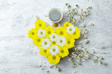 A heart shape made of bright yellow flowers with white centers, adorned with delicate white baby's breath on a rustic white wooden surface.の写真素材