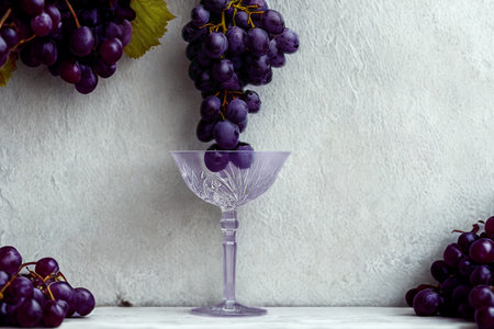 A still life featuring dark purple grapes spilling from a bunch into a decorative glass, with more grapes framing the scene.の写真素材