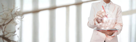 A woman in a flowing white dress stands indoors, holding a delicate bouquet of flowers, with soft light filtering through blinds.の写真素材