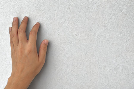 A person's hand with fingers extended gently touches a light-colored, textured wall, exploring its surface.の写真素材
