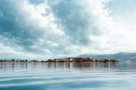 A small island village with buildings is reflected in calm, rippling water under a vast, moody sky filled with dramatic clouds.の写真素材