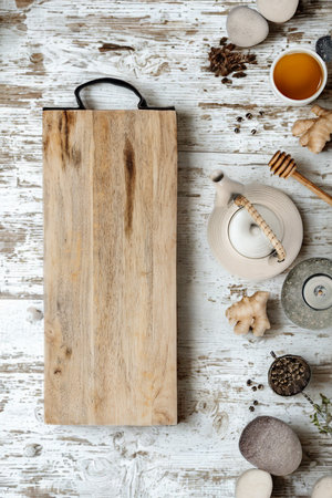 A rustic wooden cutting board sits on a distressed white surface surrounded by various spices, ginger, and a small bowl of honey.の写真素材