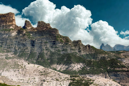 Rugged, rocky mountain peaks rise dramatically against a vibrant blue sky filled with large, fluffy white clouds.の写真素材
