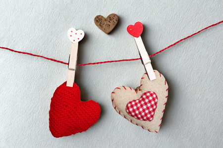 Two handmade heart-shaped decorations, one red and one patterned, are clipped to a red string with wooden clothespins against a light gray background.の写真素材