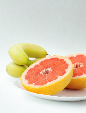 Two vibrant grapefruit halves sit on a white plate next to a bunch of bananas against a clean white background.の写真素材