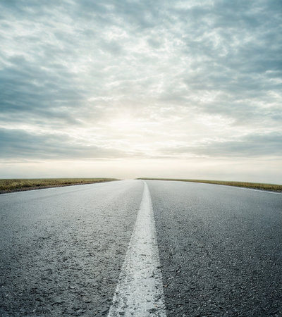 A straight, empty road stretches towards a bright horizon under a vast, cloudy sky, suggesting a journey.の写真素材