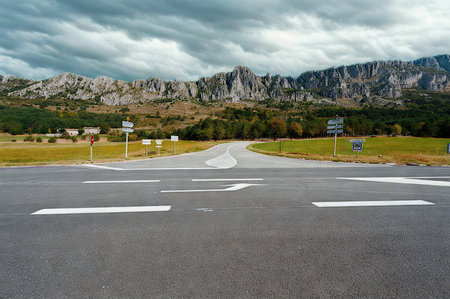 An asphalt road splits into two paths, leading towards a rugged mountain range covered in trees and dramatic clouds.の写真素材