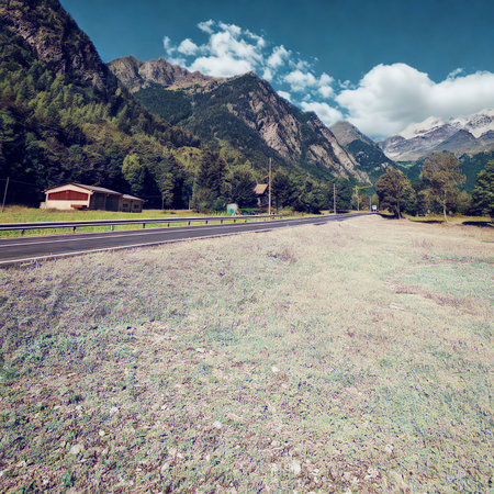 A paved road curves through a mountainous landscape with a red building nestled amongst trees under a partly cloudy sky.の写真素材