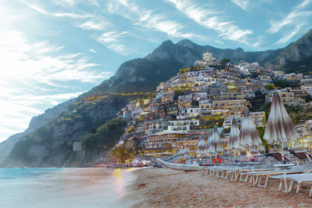 A vibrant, densely packed village cascades down a rugged mountain slope towards a serene, sunlit beach and the sparkling sea.の写真素材