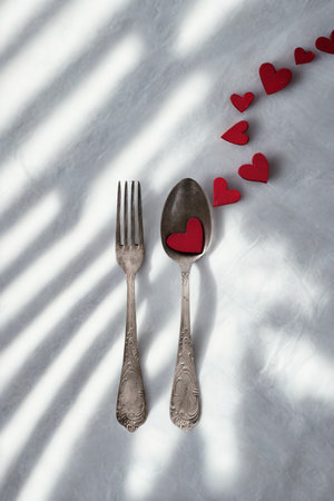 A fork and spoon are laid out on a textured surface, with a trail of red hearts leading away and dappled light creating shadows.の写真素材