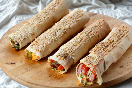 Four freshly prepared shawarma wraps, generously sprinkled with sesame seeds, are presented on a rustic wooden cutting board.の写真素材