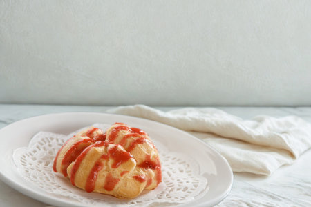 A single, golden pastry with red icing drizzled over it sits on a white plate with a lace doily.の写真素材