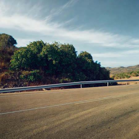 A paved road runs alongside a dry, grassy field with a dense line of trees on the other side. Hills are visible in the distance.の写真素材