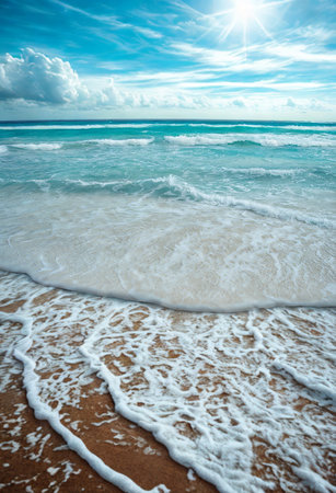 The turquoise ocean meets a sandy shore with foamy waves gently rolling in under a bright, sunlit sky with clouds.の写真素材
