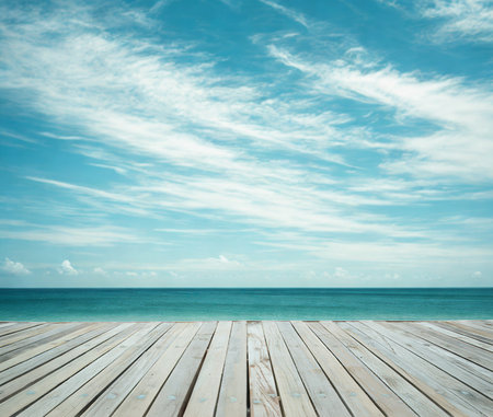 A weathered wooden boardwalk extends towards a tranquil blue ocean under a sky filled with wispy white clouds.の写真素材
