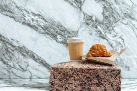 A light brown coffee beverage and a golden croissant sit on a textured block, set against a striking marble backdrop.の写真素材