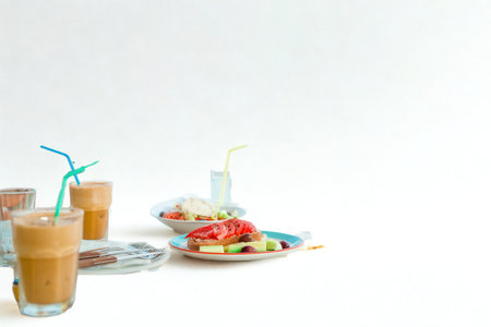 Two tall glasses of iced coffee with straws sit next to a plate of food and a bowl of fruit on a white surface.の写真素材