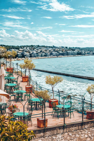 An outdoor seating area of a restaurant with tables and chairs set up along the coast, offering a view of the sea and distant buildings.の写真素材