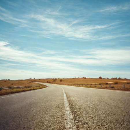 A paved road stretches into the distance through a dry, grassy landscape under a bright, cloudy sky.の写真素材