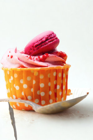 A close-up of a vibrant orange polka dot cupcake topped with swirled pink frosting and two colorful macarons.の写真素材