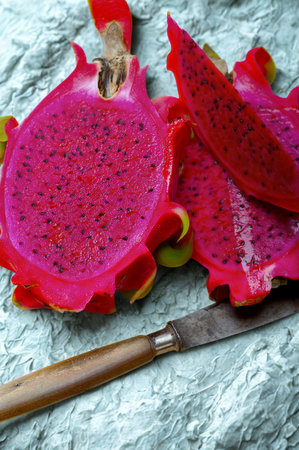 A bright pink dragon fruit is sliced in half, revealing its seeded interior. A rustic knife rests on a textured surface.の写真素材