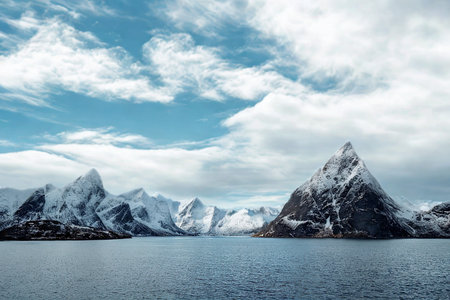 Dramatic snow-covered peaks pierce the sky above a tranquil, rippling ocean, under a dynamic cloudscape.の写真素材