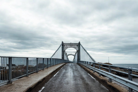 A perspective shot of a weathered suspension bridge with a wet roadway leading towards a central...