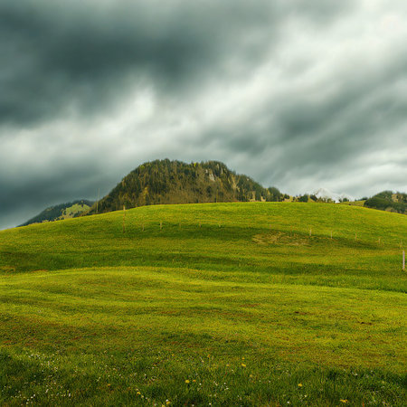 A vibrant green hillside stretches towards a rocky outcrop under a dramatic, dark, and cloudy sky, suggesting approaching weather.の写真素材