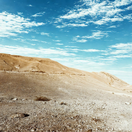 A wide, rocky, arid terrain stretches towards a distant, sun-drenched hillside under a dynamic, cloud-filled blue sky.の写真素材