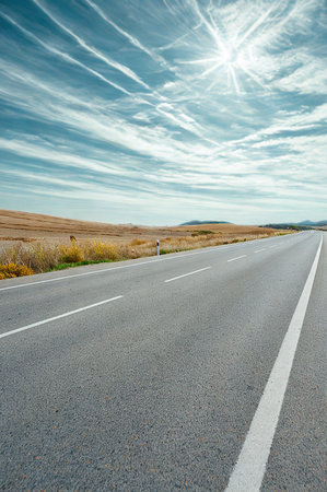 An asphalt road stretches into the distance under a bright, sunlit sky with wispy, streaking clouds.の写真素材