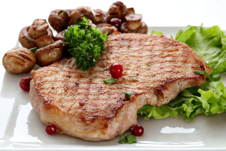 A perfectly cooked pork chop served with sautéed mushrooms, fresh lettuce, and garnished with peppercorns on a white plate.の写真素材