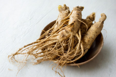 A collection of natural, unprocessed ginseng roots with delicate fibers, displayed in a simple wooden bowl on a textured surface.の写真素材