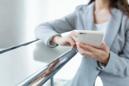 A woman in a professional suit holds a smartphone, her hand resting on a metallic railing, suggesting a moment of pause or connection.の写真素材