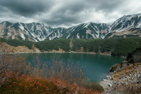 A tranquil lake reflects the dramatic sky and surrounding mountains, with vibrant autumn colors in the foreground and snow on the peaks.の写真素材