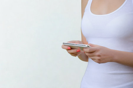 A close-up shot of a woman's hands holding a silver smartphone against a white background. She is wearing a white tank top.の写真素材